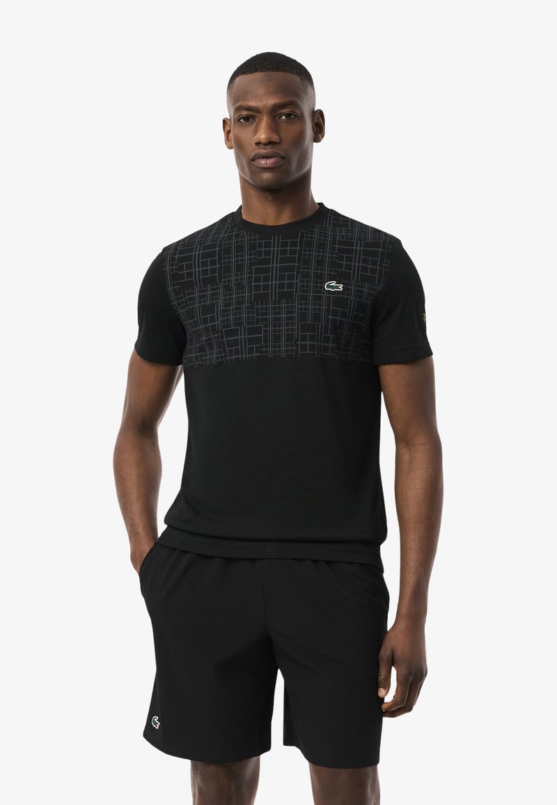 Un uomo sta in piedi con sicurezza indossando una t-shirt nera con una parte superiore a fantasia e pantaloni corti neri, con un piccolo logo verde Lacoste.