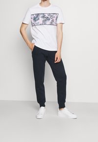 Mies, który ma na sobie białe trampki, granatowe dresy i białą koszulkę z różowo-niebieskim wzorem liści na piersi, stoi na jednolitym tle.