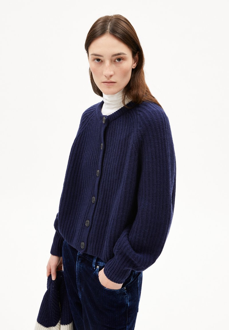 Navy blue knitted cardigan with ribbed texture, round neckline, and large buttons. Model wears it over a white turtleneck, paired with dark pants.