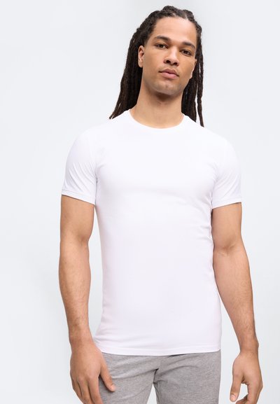 Άνδρας με μακριές πλεξούδες dreadlocks, φορώντας εφαρμοστό απλό λευκό κοντομάνικο πουκάμισο και ανοιχτό γκρι παντελόνι, μπροστά σε απλό φόντο.