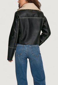 Schwarze Leder-Cropped-Jacke mit einem beigen Lammfellkragen, sichtbaren Nähten und leicht ausgestellten Ärmeln. Blaue Jeans runden den Look ab.