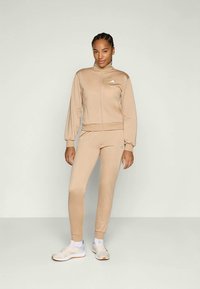 Bež set sportskog trenerke koji se sastoji od jakne na zip s rebrastim manžetama i odgovarajućih suženih hlača, uparenih s bijelim tenisicama.