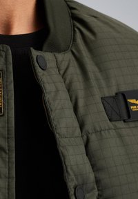 Zielona kurtka z wypełnieniem w wzór kratę. Posiada prążkowany kołnierz, czarne guziki oraz naszywkę z logo z boku. Teksturalny materiał.