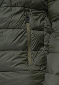 Maslinasto zeleni puffer jakna s kvadriranom teksturom, koja sadrži džep sa zatvaračem od istog materijala i crne kopče.