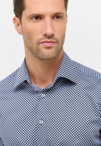 Man met lichte stoppels in een blauw gestreept overhemd, die iets opzij kijkt tegen een witte achtergrond.
