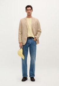 Beige linnen blazer over een lichtgele t-shirt, blauwe straight-leg jeans en donkere schoenen. Houdt een gele pet vast. Neutrale achtergrond.