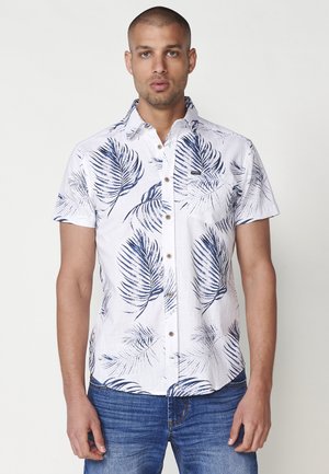 Camisa de manga corta en tela blanca con estampado de hojas de palmera en azul marino, con botón en el frente y bolsillo en el pecho. Diseño casual con un corte holgado.