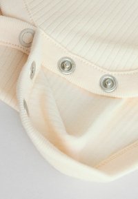 Tessuto a coste color crema con una superficie liscia, dotato di bottoni a pressione metallici sul colletto per la chiusura.