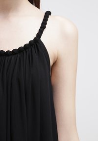 Black sleeveless dress with elasticized neckline, featuring gathered fabric and textured straps made of rounded accents. Soft, flowing texture.