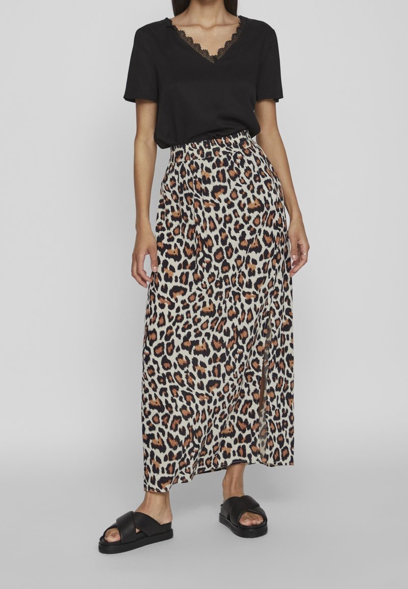 Femme portant un haut noir à manches courtes avec un col en dentelle, une jupe longue taille haute à imprimé léopard avec une fente à l'avant, et des sandales noires à lanières croisées.