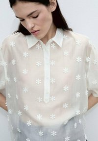 Vit broderad blus med korta ärmar, med blommotiv, knäppning fram och en genomskinlig textur.
