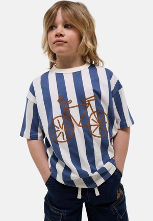 Enfant blond portant une chemise rayée verticale bleu et blanc avec un vélo brun brodé, mains dans les poches d’un pantalon cargo bleu foncé.