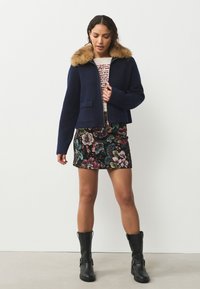 Giacca corta blu navy con collo in pelliccia sintetica, tessuto strutturato, due tasche frontali. Abbinata a una minigonna ricamata a fiori e stivaletti neri alla caviglia.