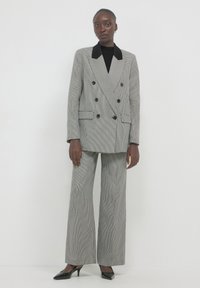 Femme debout portant un blazer croisé noir et blanc à motif pied-de-coq et un pantalon large assorti, avec des talons noirs.