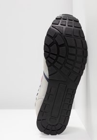 Semelle d'une chaussure de sport grise avec une semelle extérieure en caoutchouc noire à motifs, inclinée verticalement contre un coin blanc.
