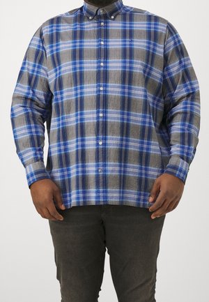 Homme portant une chemise à carreaux bleue, grise et blanche boutonnée avec un pantalon foncé, debout devant un fond uni.