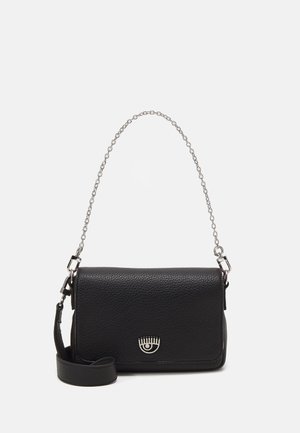 Sac bandoulière en cuir noir avec logo argenté en forme d'œil, chaîne argentée amovible et bretelles en cuir sur fond blanc.
