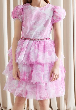 Fille portant une robe fleurie rose avec des manches bouffantes, une jupe à volants et une ceinture ornée de bijoux, debout devant des rideaux beiges.