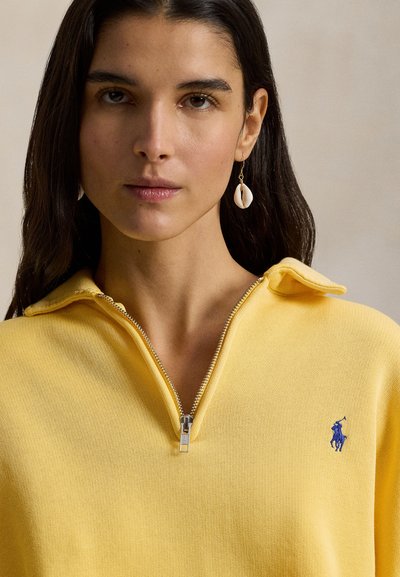 Κίτρινο φούτερ με φερμουάρ σε ριμπ υφή με μικρό μπλε λογότυπο. Διαθέτει γιακά που στέκεται και ασημένια λεπτομέρεια φερμουάρ.