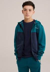 Huvtröja i teal och marinblå, med dragkedja, sidofickor och en mjuk textur, tillsammans med en marinblå crew-neck-tröja och matchande byxor.