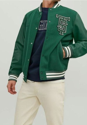 Homme portant une veste varsity verte avec poignets et col rayés blancs, sweat-shirt bleu marine et pantalon beige clair, main dans la poche.