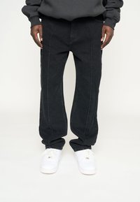  Sorte straight-leg bukser bæres med hvide sneakers og en mørk sweatshirt, hænderne hvilende ved siderne mod en lys baggrund.