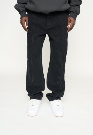Black denim pants with a relaxed fit, featuring front seams, deep pockets, and rolled cuffs, paired with white sneakers.
