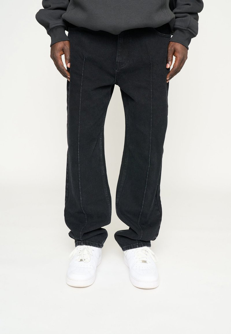  Sorte straight-leg bukser bæres med hvide sneakers og en mørk sweatshirt, hænderne hvilende ved siderne mod en lys baggrund.