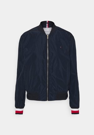 Veste bomber bleu marine avec fermeture éclair devant, col et poignets côtelés avec des rayures rouges, blanches et noires, et petit logo sur la poitrine.