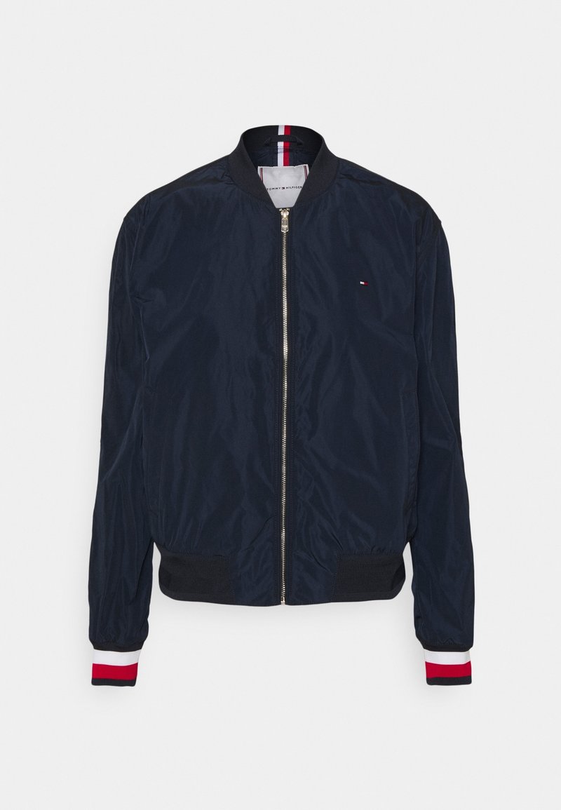 Veste bomber bleu marine avec fermeture éclair devant, col et poignets côtelés avec des rayures rouges, blanches et noires, et petit logo sur la poitrine.