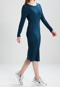 Abito lungo a maniche lunghe di color teal, con dettagli di cucitura curva; lunghezza al ginocchio. Abbinato a sneakers argentate metallizzate. Tessuto liscio, forma aderente.