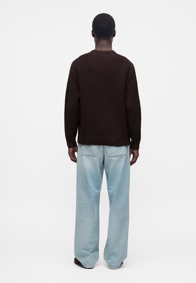 Personne debout, dos à la caméra, portant un pull marron foncé et un jean large bleu clair sur fond blanc.