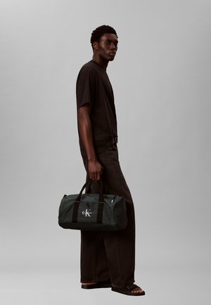 Uomo con t-shirt nera, jeans neri larghi e sandali tiene una borsa duffel Calvin Klein verde scuro su sfondo grigio.