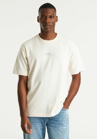 T-shirt crema a maniche corte con scollatura rotonda e un sottile logo blu sul petto, abbinato a jeans azzurri chiari strappati.