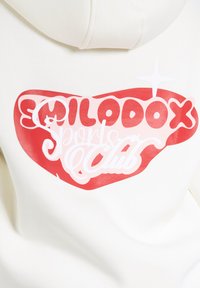 Felpa color crema con un grande grafico di un cuore rosso e la scritta "SMILODOX Sports Club" in grassetto bianco e rosa.