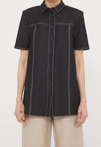 Camicia nera a maniche corte con bottoni, con cuciture verticali e orizzontali bianche, indossata con pantaloni beige.