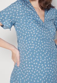 Blaues Blumenkleid mit kurzen Ärmeln, das mit kleinen weißen Blumen verziert ist, einem V-Ausschnitt und einer taillierten Taille. Glatte Stofftextur.