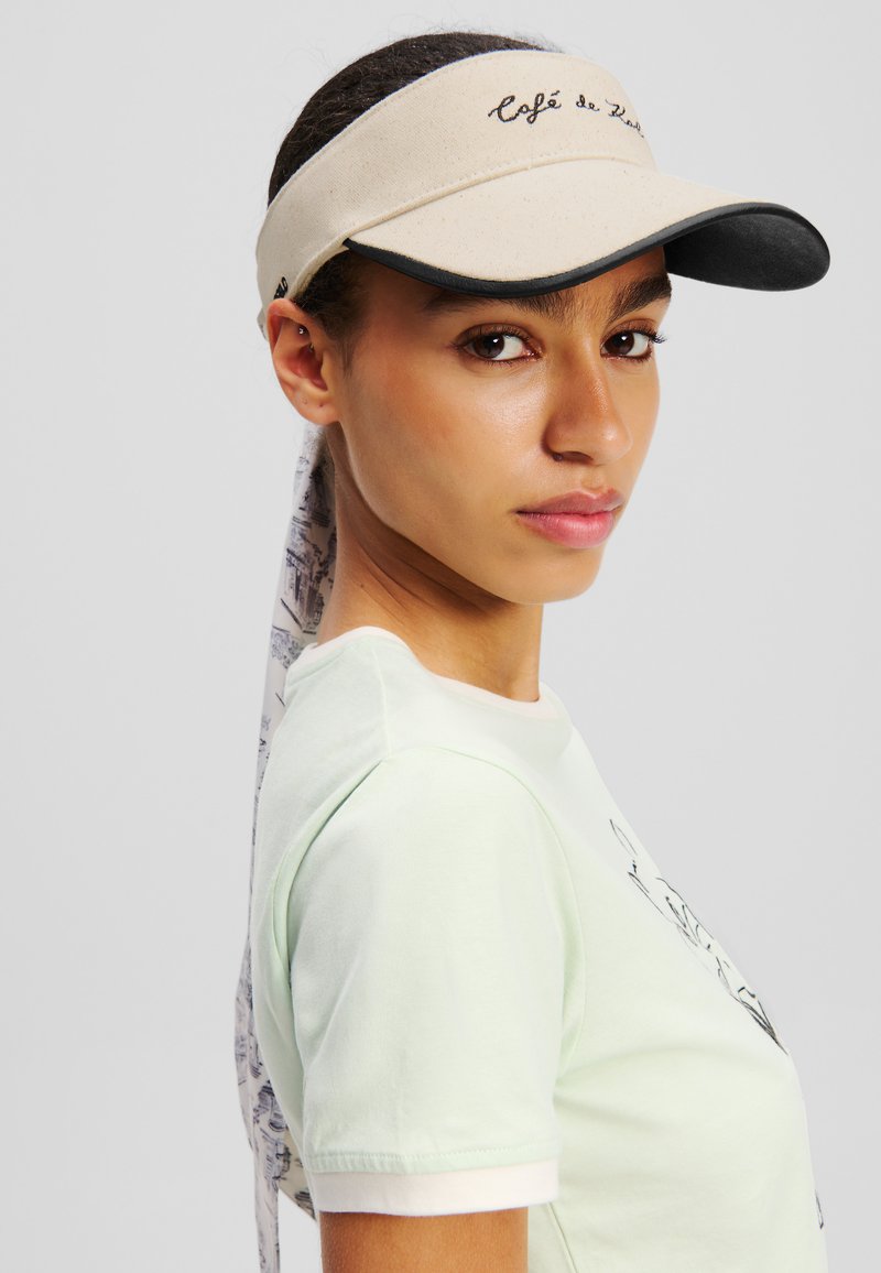 Beige visir med sort skygge, broderet "Café de Zel", parret med en lys grøn t-shirt. Visiret har et mønstrede tørklæde bagpå.