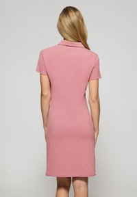 Sukienka polo w miękkim różowym materiale, z krótkimi rękawami, dopasowaną sylwetką i prostym dołem. Gładka tekstura z klasycznym kołnierzykiem.