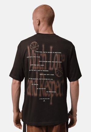 Persoon met kort haar draagt een zwart Jordan T-shirt met inspirerende basketbalthematekst op de achterkant.