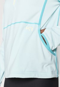 Ανοιχτό μπλε μακρυμάνικη μπλούζα με στρογγυλεμένη κάτω ραφή, σ ε αντίθεση ραφές σε τιρκουάζ, λογότυπο και πλαϊνή τσέπη για αποθήκευση. Λείο, αθλητικό ύφασμα.