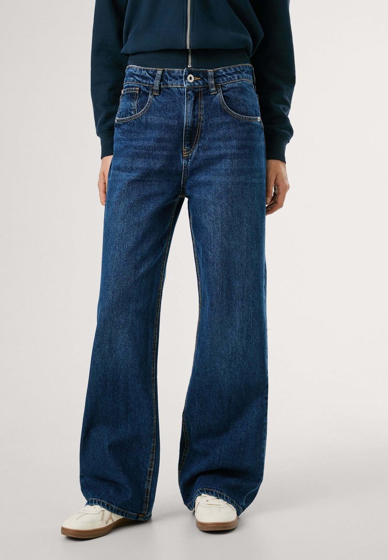 Persoon in donkerblauwe wijde jeans, witte sneakers en een marineblauw langemouwtop met een ritsdetail.