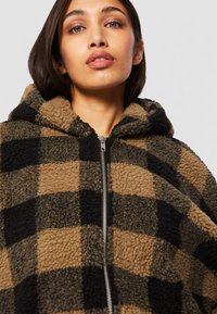 Braun und schwarz kariertes Fleece-Hoodie mit Reißverschluss, ausgestattet mit einer plüschigen Textur und einer geräumigen Kapuze. Das gemusterte Material sorgt für visuelles Interesse.