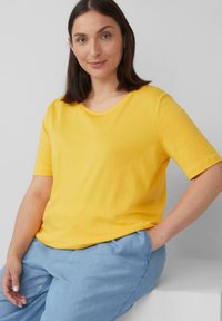 Frau mit schulterlangem braunem Haar, die ein gelbes kurzärmliges Shirt und hellblaue Hosen trägt, sitzt mit einer Hand in der Tasche vor einem grauen Hintergrund.