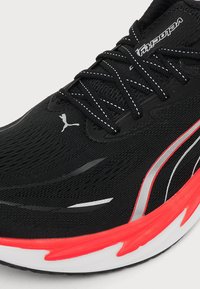 Chaussure de sport noire avec semelle rouge et blanche, lacets noirs avec surpiqûres blanches, logo Puma et bandes latérales argentées et rouges.