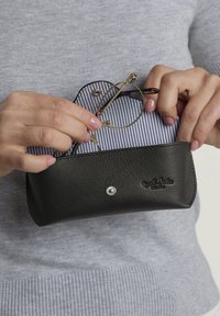 Étui à lunettes en cuir noir avec un fermoir à bouton-pression argenté, contenant des lunettes rondes contre un tissu doublé à rayures blanches et bleu marine.