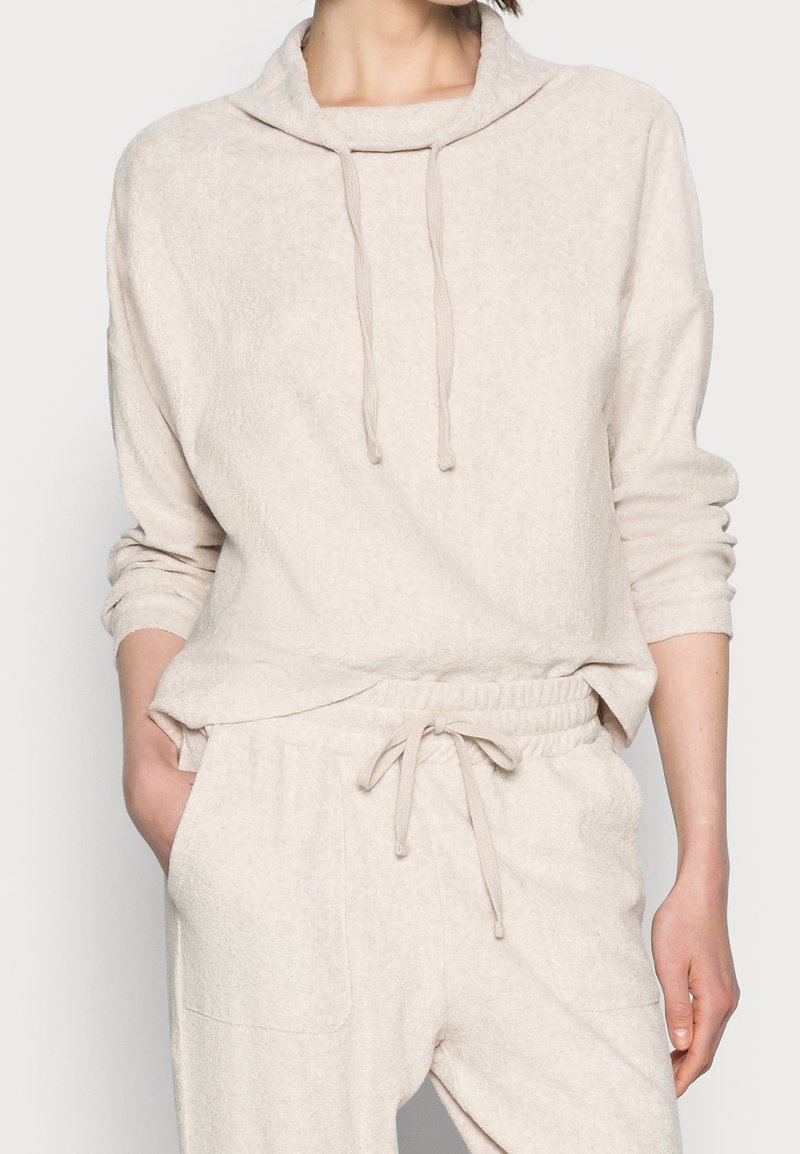 Femme portant un sweat à capuche en tissu doux beige et un pantalon à cordon assorti, la main gauche dans la poche, sur un fond uni.