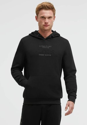 Man with short hair wearing a black hooded sweatshirt with front pocket and printed text "urban trail running CAMP DAVID" on chest.