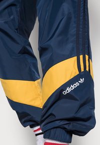 Navy trackbyxor med gula detaljer, med en Adidas-logotyp. Materialet är glänsande och slätt, med elastiska ärmslut och vertikala ränder.