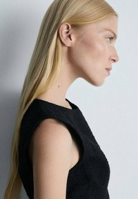 Svart texturerad ärmlös topp med rund halsringning, sedd på en modell med rakt blont hår, som framhäver tygets detaljer och struktur.
